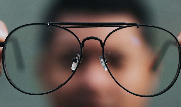 A Black man is out of focus in the background, he holds out his glasses, which are clear in the foreground