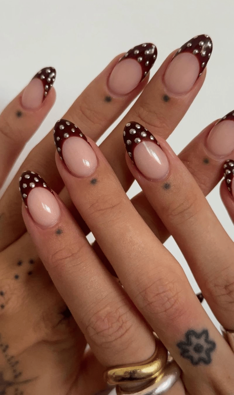 Almond nude nails featuring burgundy French tips embellished with small silver studs.