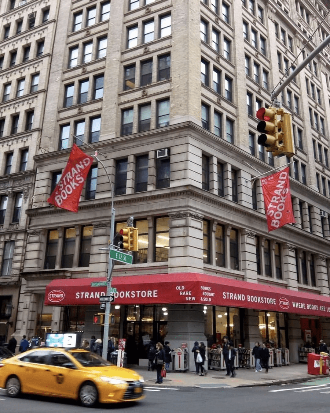 Street view of the Strand Bookstore in NYC with its iconic red awning, book displays, and a yellow taxi passing by.
