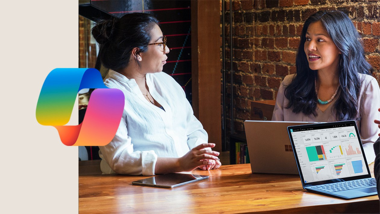 Two women sit at a wooden table with a laptop, tablet, and charts on the screen, discussing work; a colorful abstract logo is on the left side of the image.