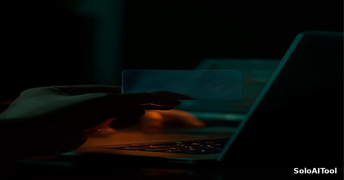 A moody close-up of a hand holding a plain credit card beside a glowing laptop on a dark desk in green and amber tones