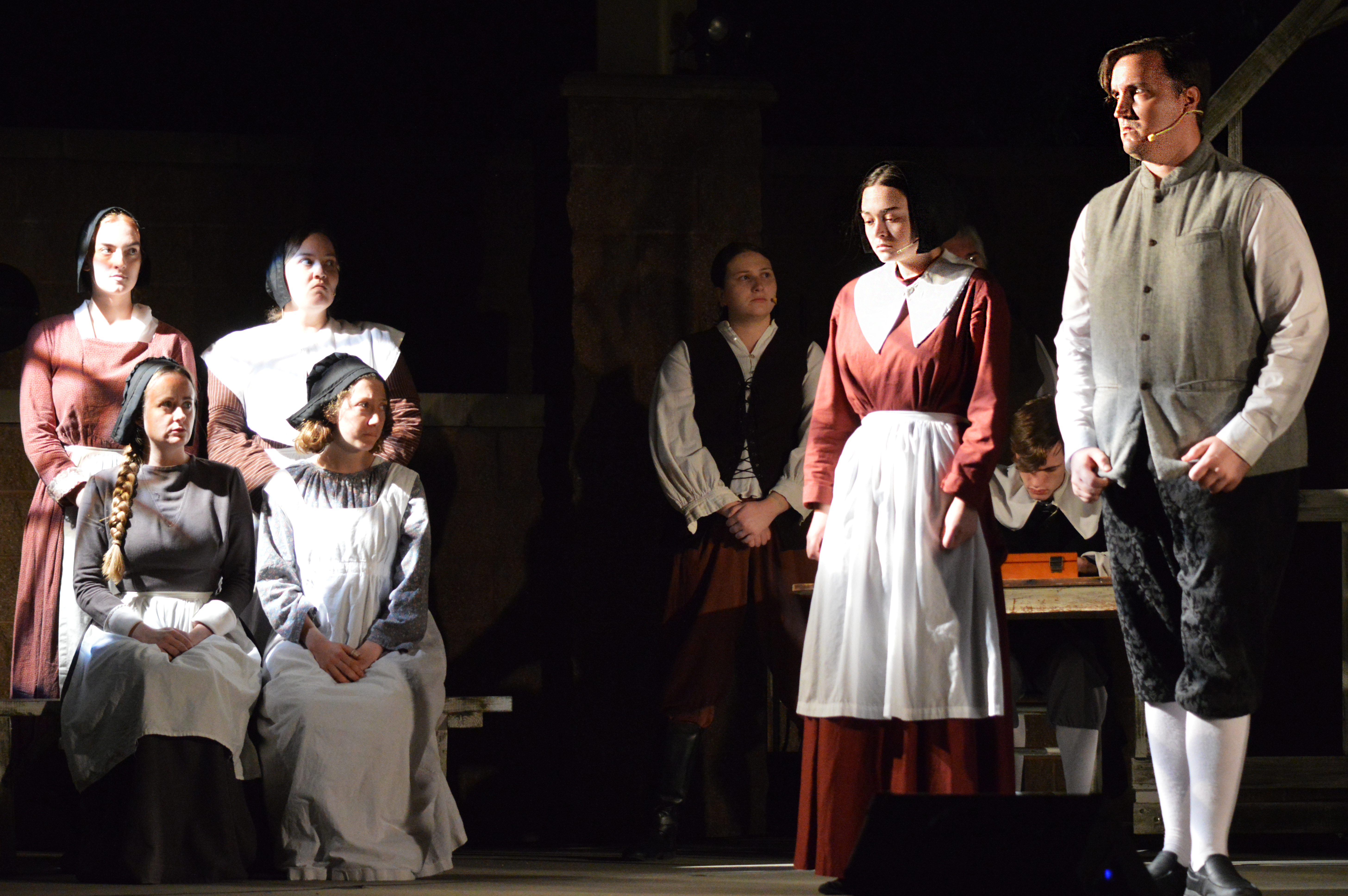 About eight theater actors stand onstage dressed in old-fashioned clothing.