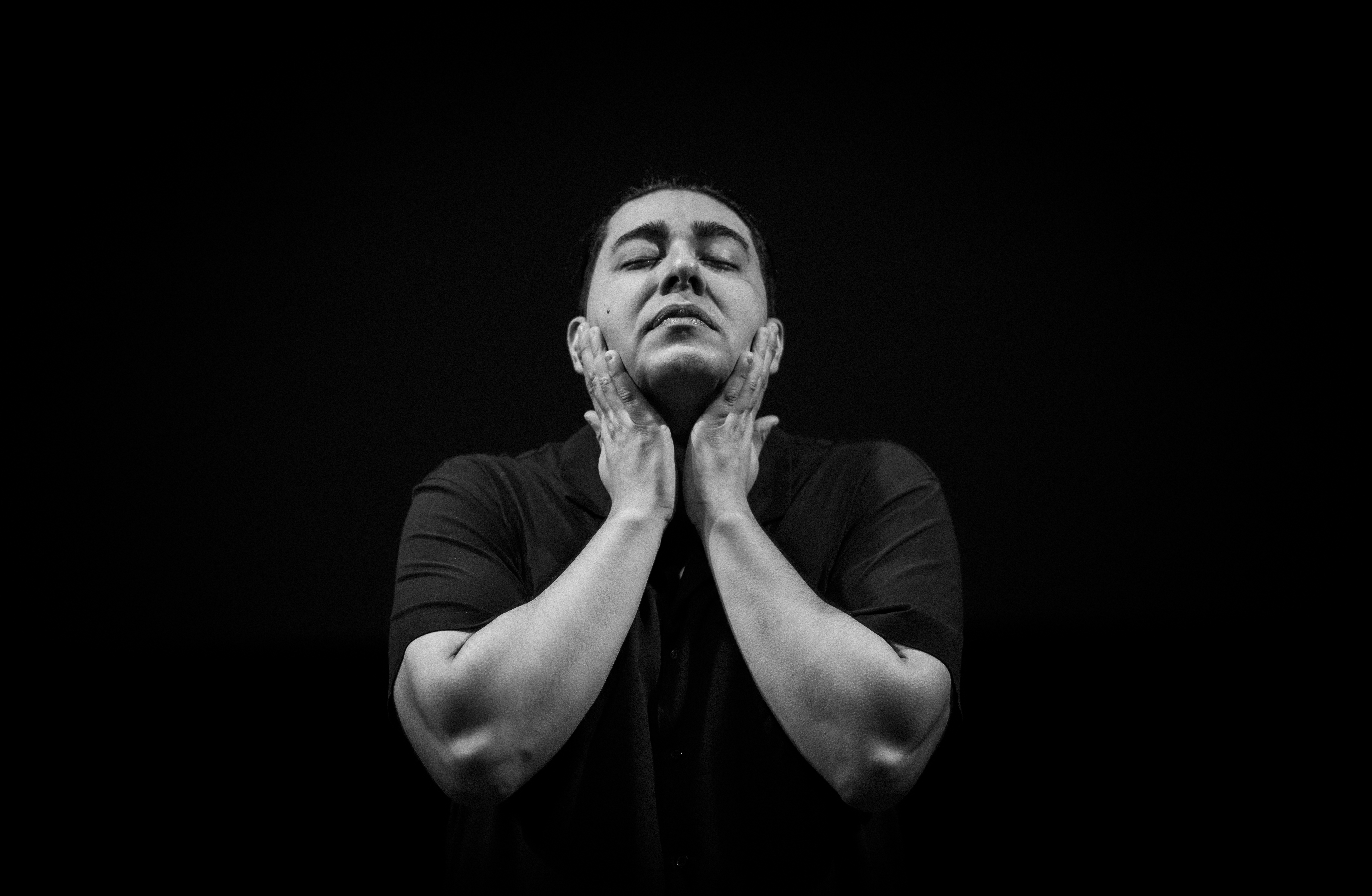 A man holds his chin, his eyes closed, facing the camera with a dark background.