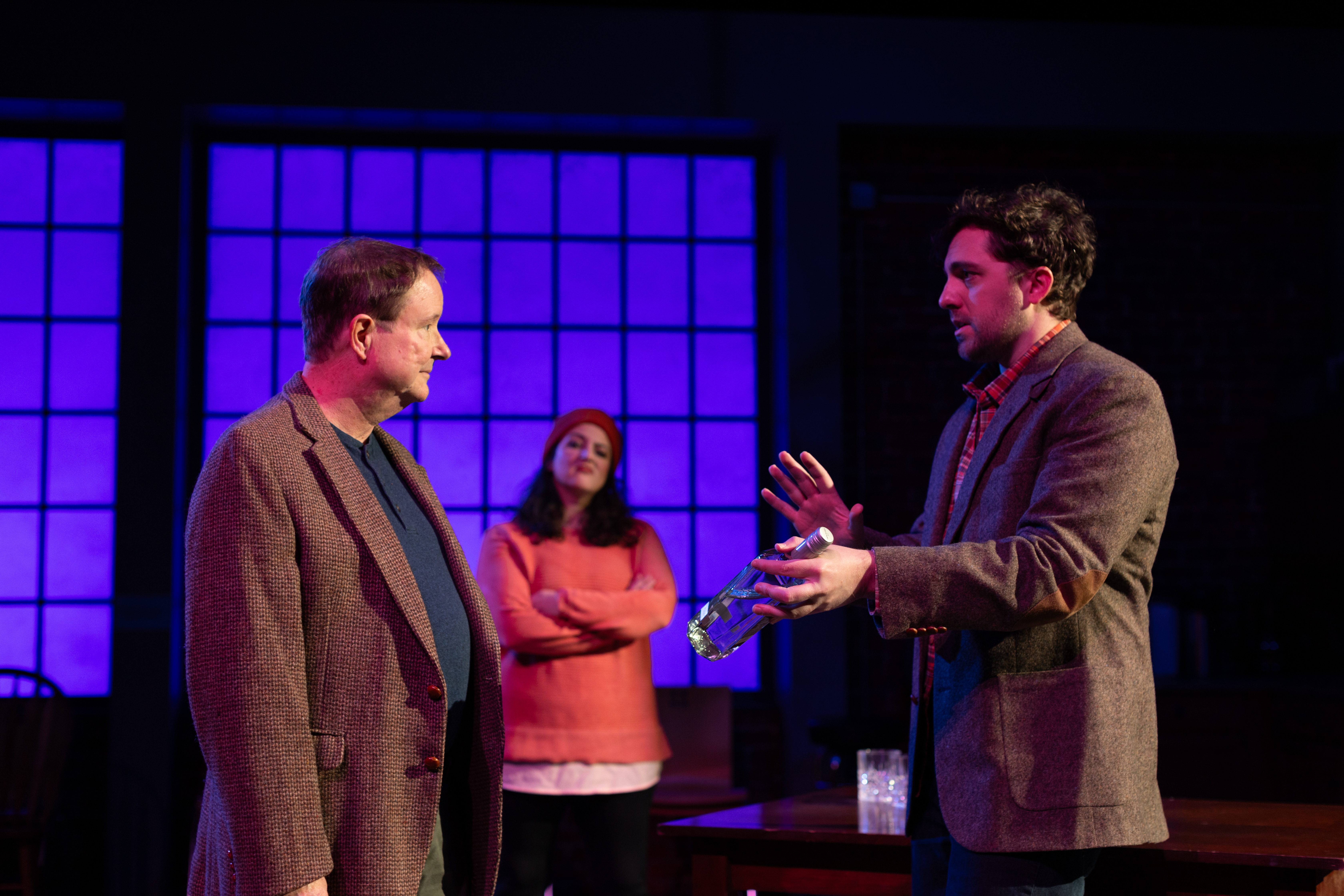 Two male actors talk to one another onstage with a female actor in the background, wit her arms crossed.