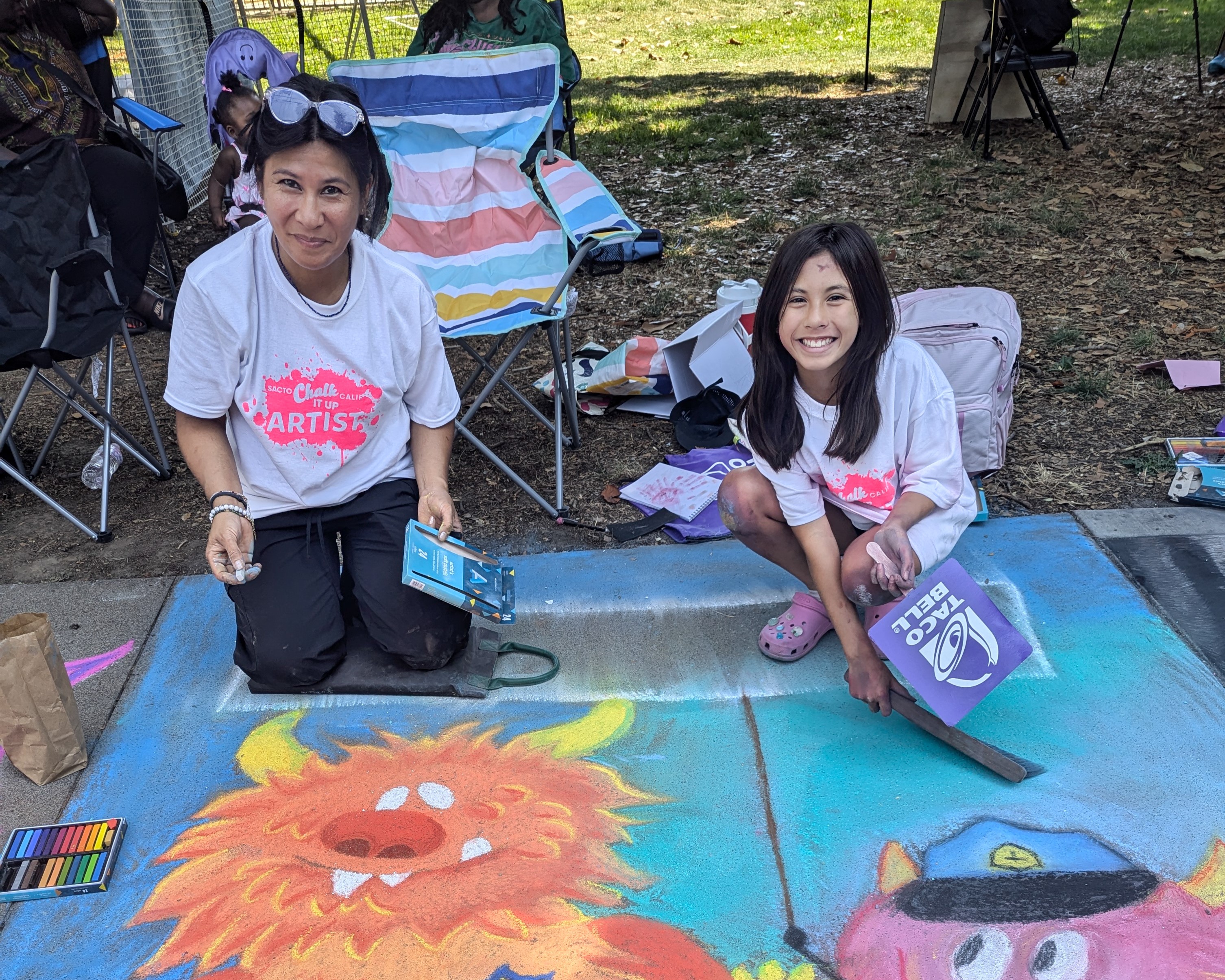 Two people crouch over chalk art with pieces of chalk in hand.