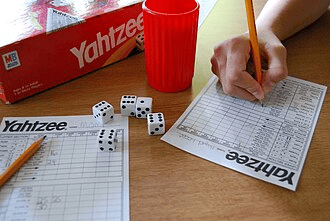 This is an image of the Yahtzee game, cup, dice, and score sheets. A hand is filling out one of the sheets. The image is sourced from Wikipedia. 