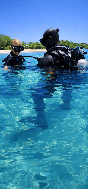 Two scuba divers wearing gear in clear shallow water near a sandy beach