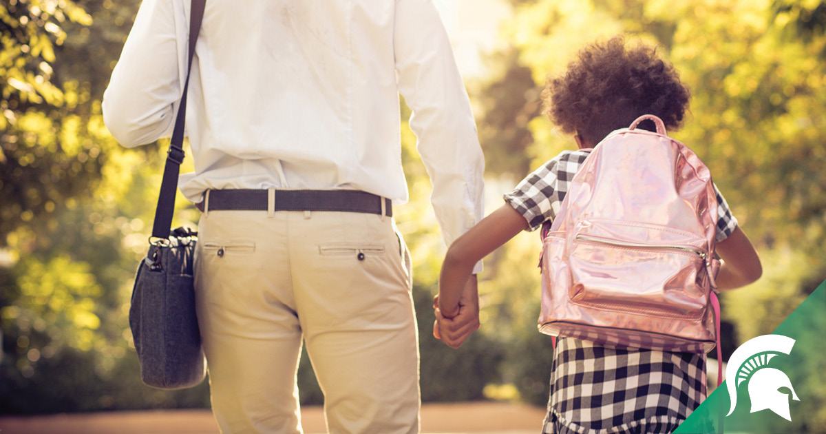Seen walking away from the camera, an adult in professional dress carrying a laptop bag holds the hand of a small child in a black and white checked dress with a shiny pink backpack.