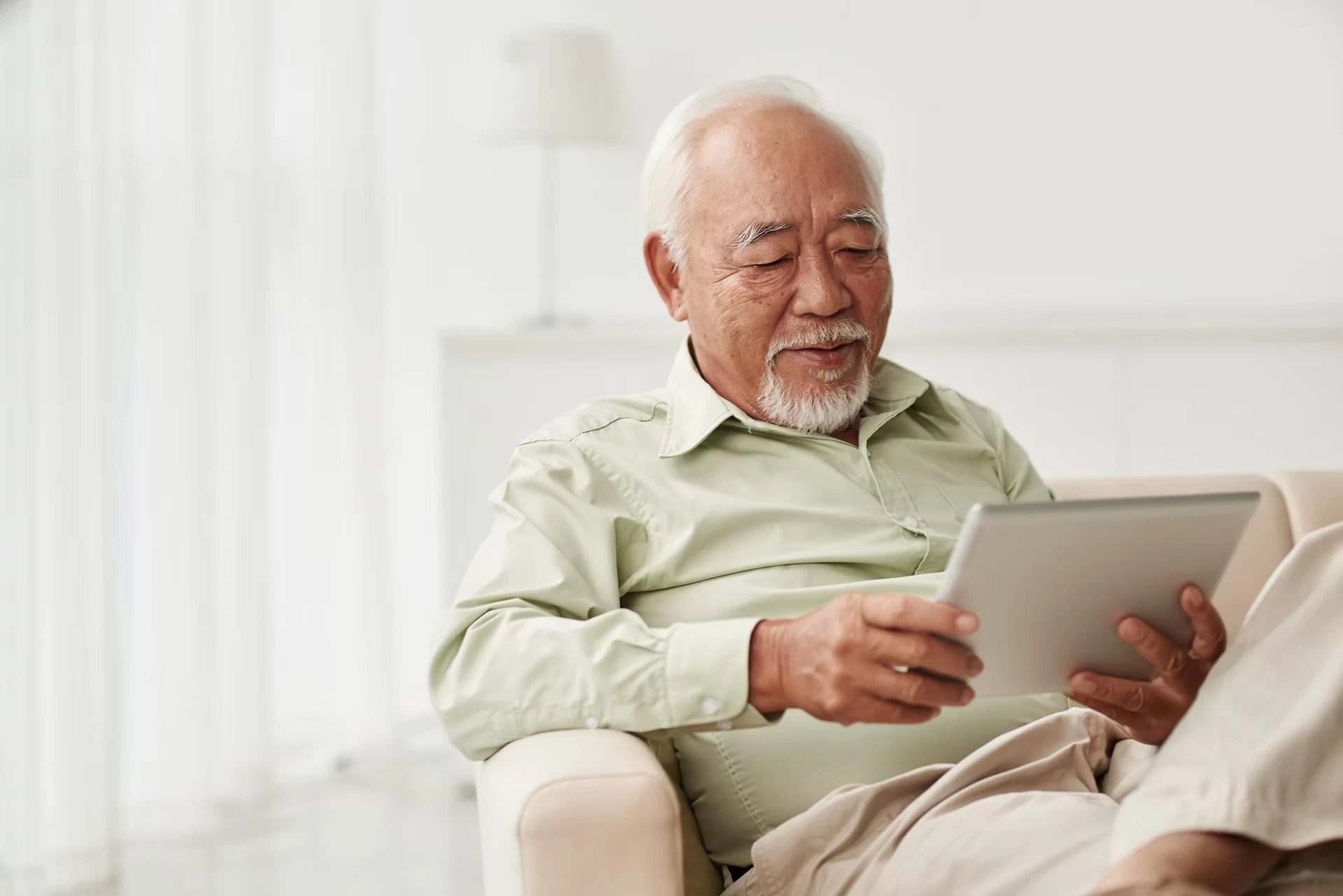 An older man using a tablet while sitting on a white couch.