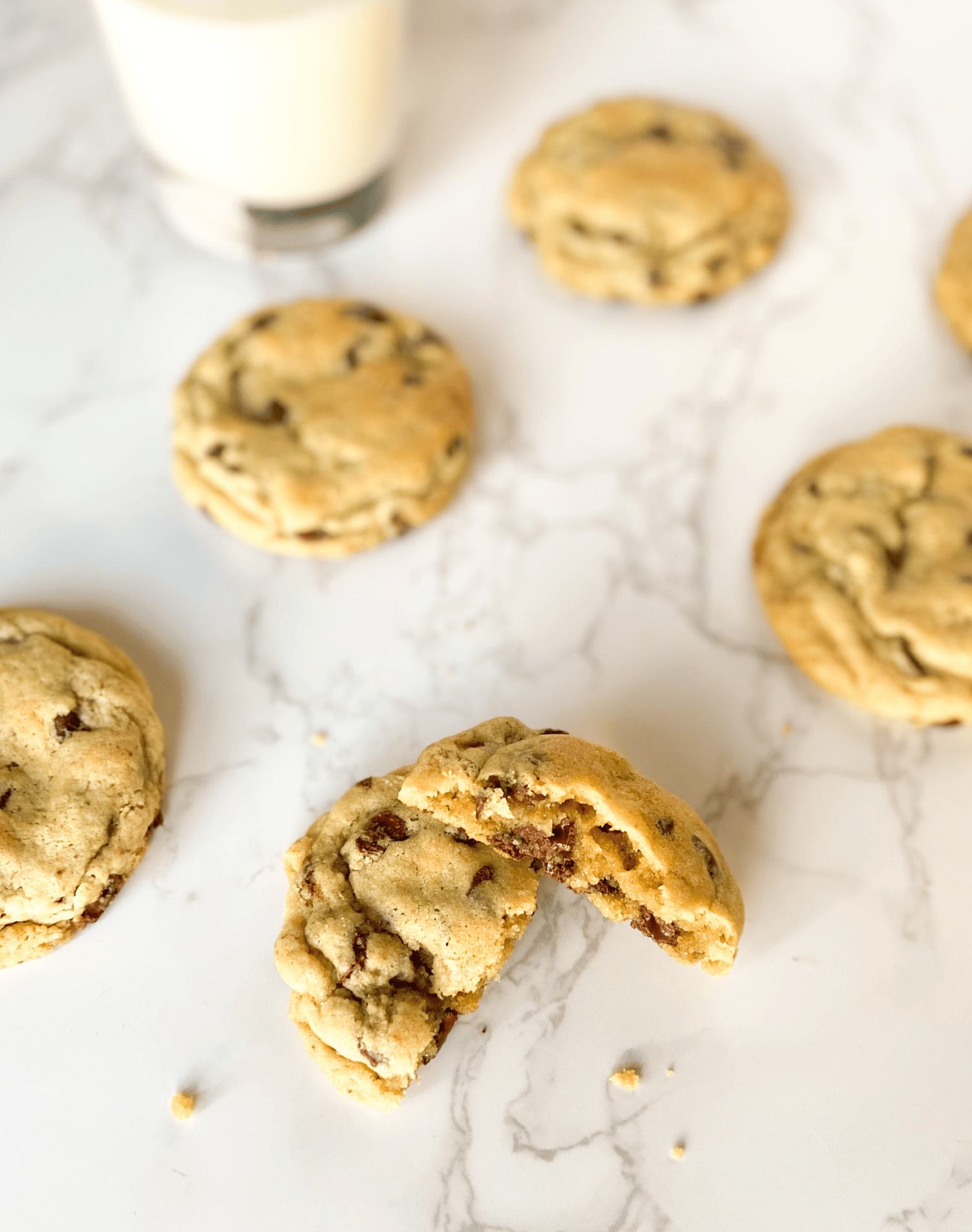 A broken chocolate chip cookie with other chocolate chip cookies. A glass of milk in the background.