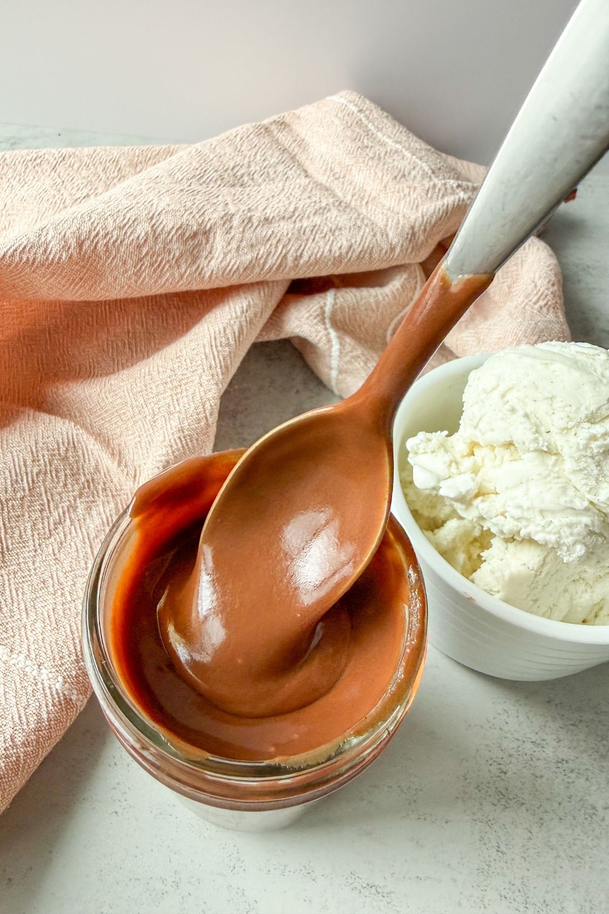 Spoonful of hot fudge coming out of a jar of hot fudge. A bowl of vanilla ice cream to the side with a hand towel in the background.