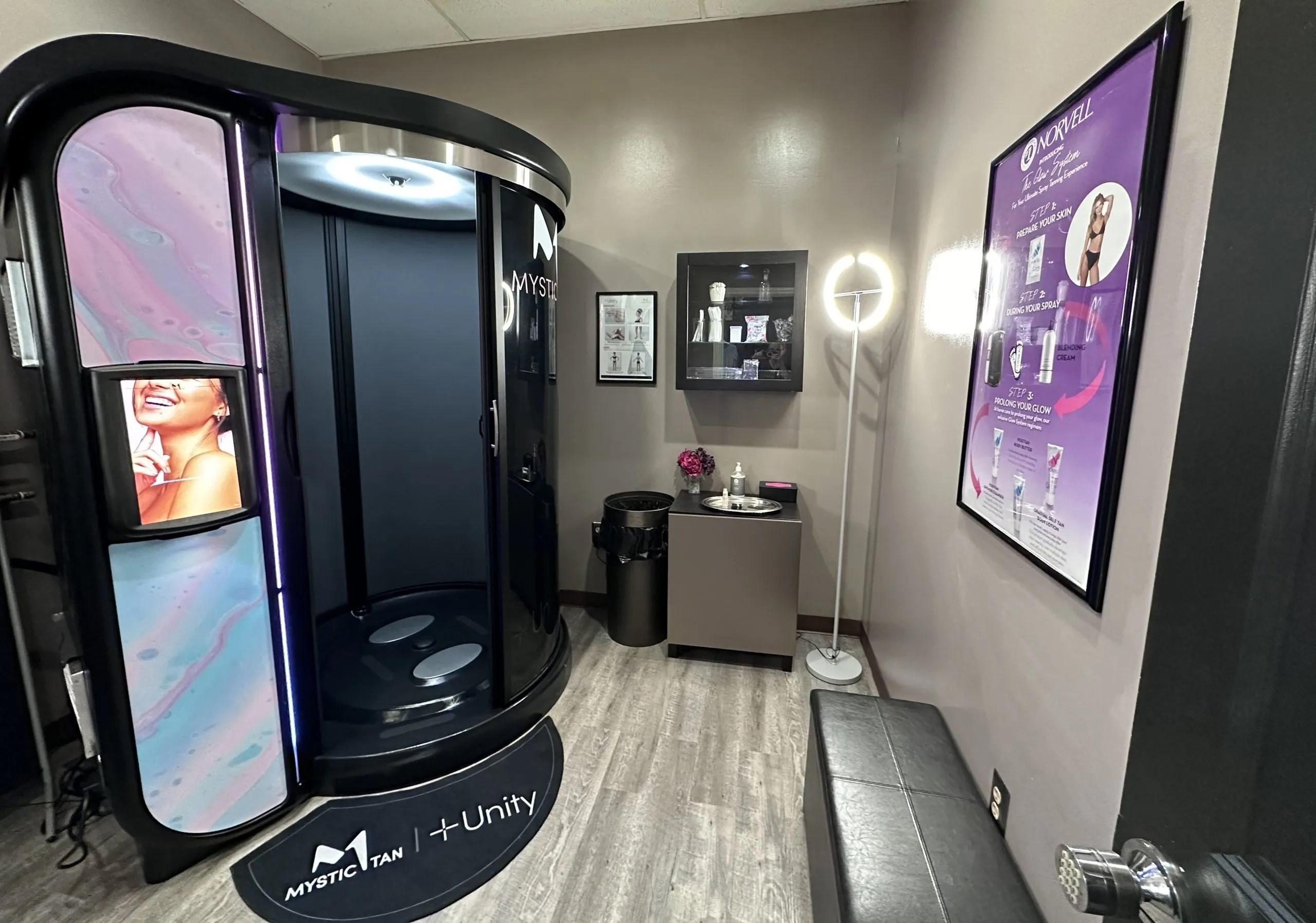 SprayChic Airbrush Tanning room featuring a Mystic Tan spray tanning booth with ‘Unity’ branding, a well-organized shelf with tanning products, and informational posters on the wall. The room is modern and inviting, with a circular LED light, a small cabinet adorned with flowers, and a comfortable bench for clients.