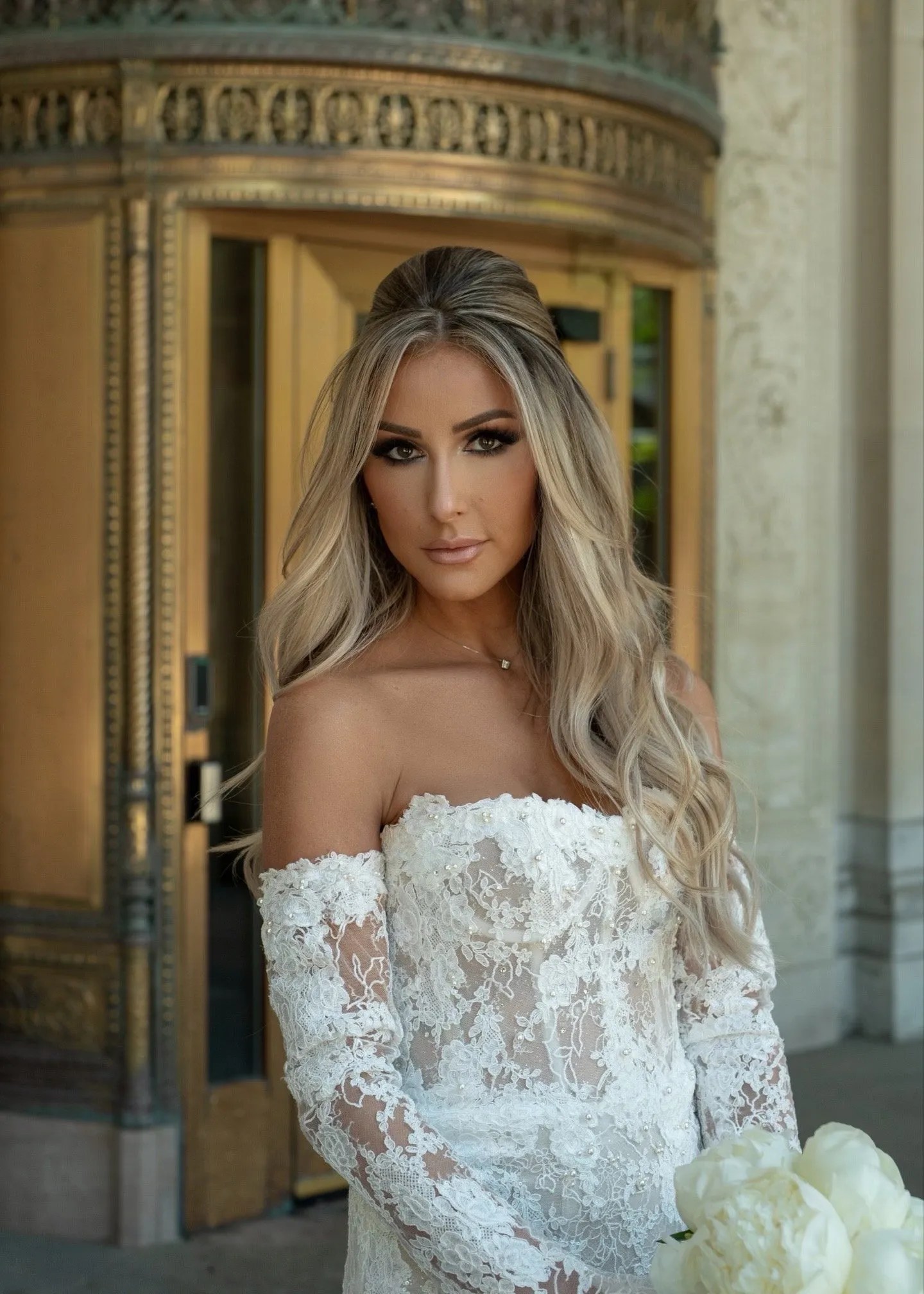 A bride with long, wavy blonde hair, wearing an off-the-shoulder lace wedding gown, posing outdoors in front of an ornate building entrance. Her makeup is elegant and subtle, complementing her flawless, sun-kissed skin achieved with a SprayChic airbrush tan. She holds a bouquet of white flowers, looking confidently at the camera.