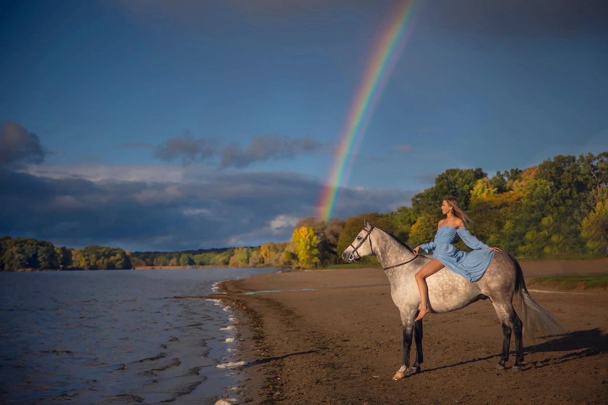 A serene scene featuring a woman with a beautiful spray tan in a flowing light blue off-shoulder dress sitting gracefully on a gray horse by the edge of a tranquil lake. A vibrant rainbow arcs across the sky, contrasting with the dark storm clouds and golden autumn foliage in the background. The woman gazes thoughtfully into the distance, exuding a sense of calm and connection with nature.