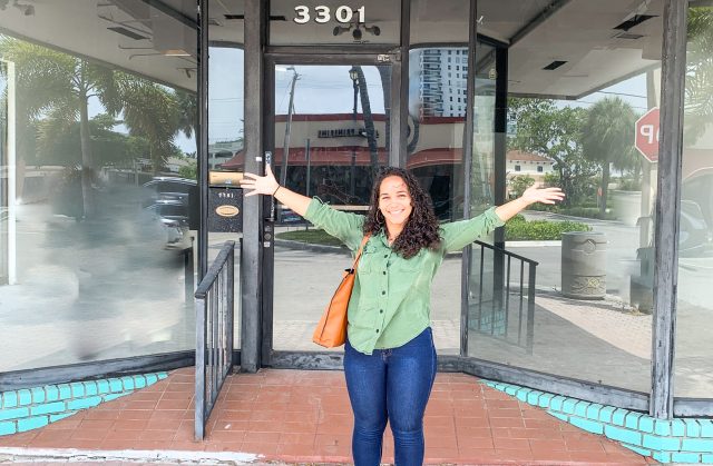 Megan Wilson, owner, standing with arms outstretched in front of the glass entrance to Spud's Coffee in Fort Lauderdale, Florida.