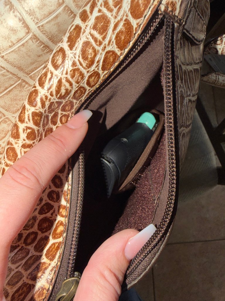 A close-up of a hand reaching into a zipped compartment of a handbag, revealing a black item partially hidden inside.