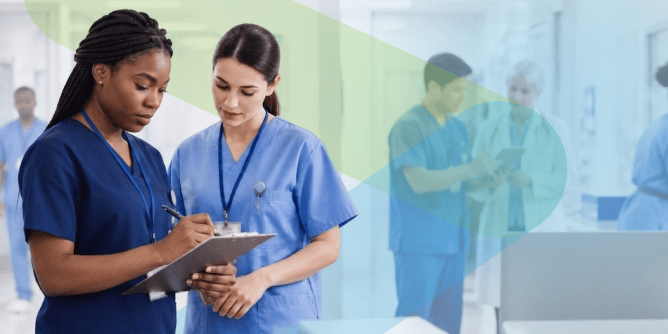 Two nurses in blue scrubs review notes on a clipboard in a hospital hallway while other healthcare staff work in the background.
