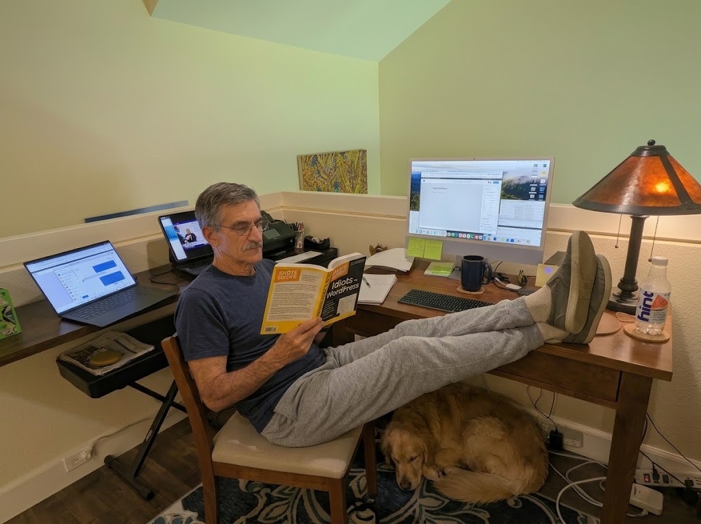 An elderly man reading a book while seated at a desk, with his feet propped up. Two computers are visible on the desk, and a golden retriever is lying on the floor nearby.