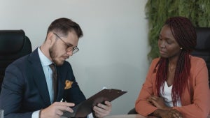A man in a suit reviewing a clipboard while sitting across from a woman with braids, both engaged in conversation in a professional setting.