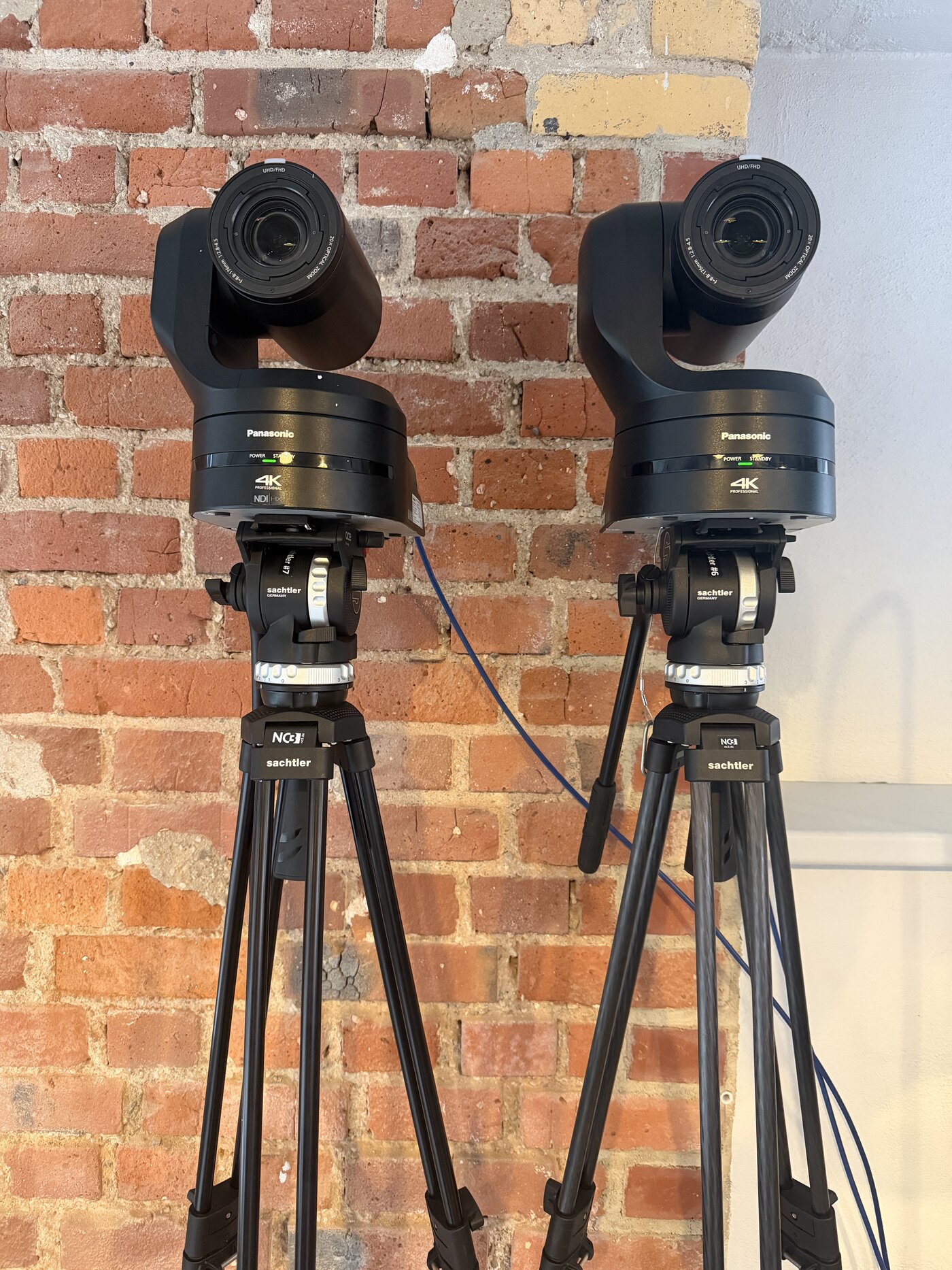 Zwei Panasonic PTZ 4K Kameras auf Sachtler Stativen mit NC3-Logo vor Backsteinwand