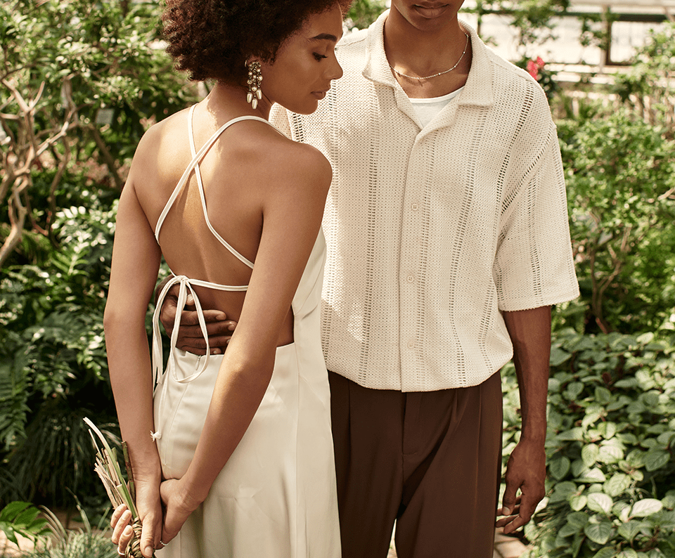 woman with white open back dress turning side and man wearing white shirt and brown pants in garden