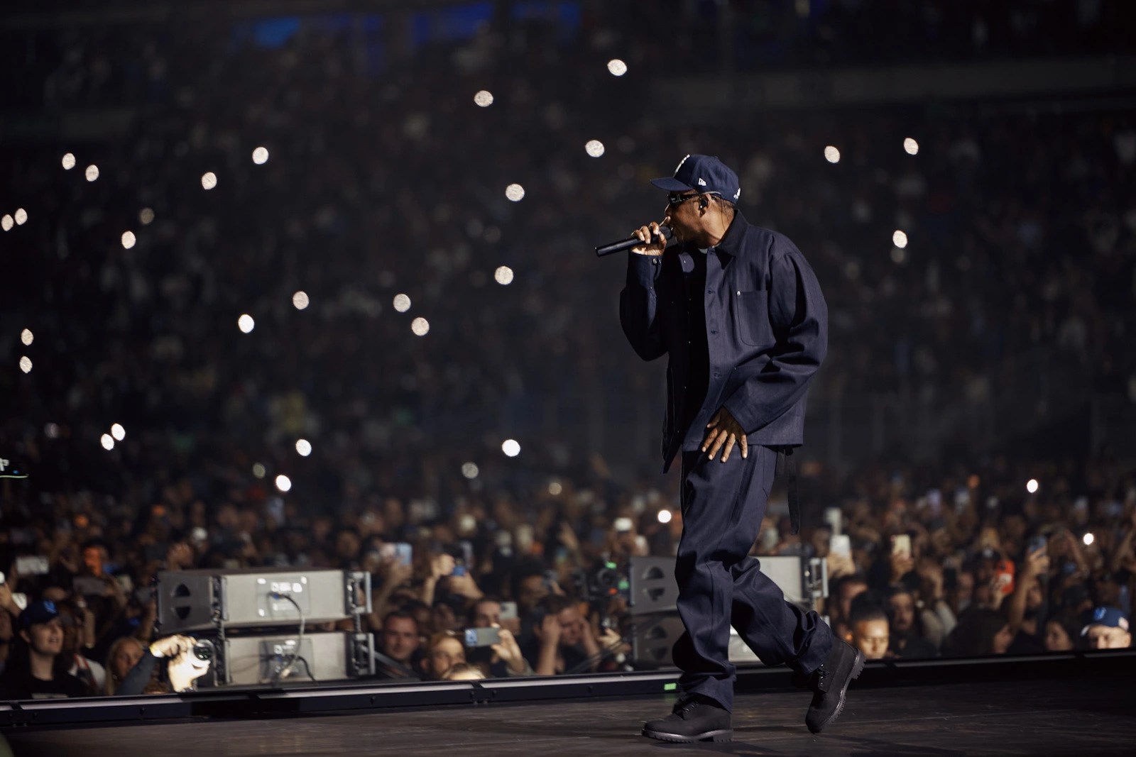 Jay-Z performing on stage in a dark outfit and cap, holding a microphone as a large crowd watches with phone lights raised.