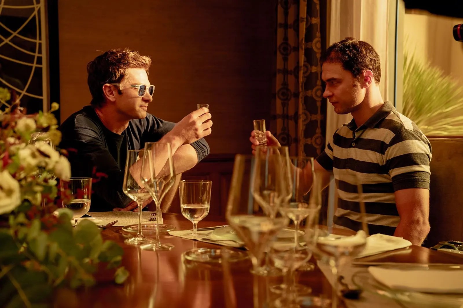 Andrew Garfield as Sam Altman raises a glass at a table alongside Yura Borisov as Ilya Sutskever in NEON’s upcoming film, Artificial.