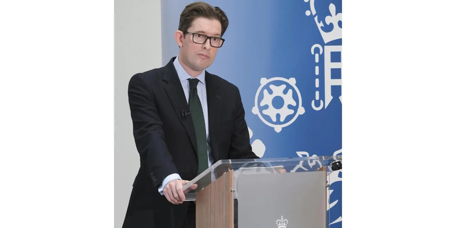 MI5 Director General Sir Ken McCallum stands behind a lectern during a speech, with a blue government security backdrop behind him.