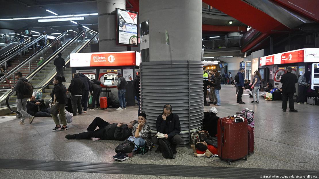 Estación de tren, con gente sentada o tumbada, y otra caminando.