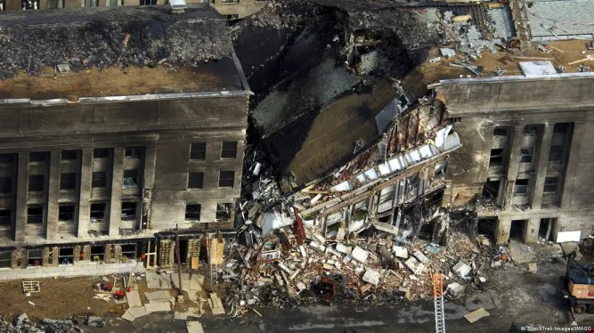 Aerial photo of a section of a long, tall building, badly damaged and smoke-blackened. The central section has collapsed.