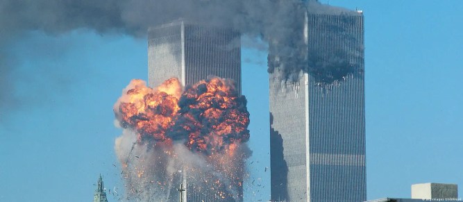 The twin towers of the World Trade Center against a bright blue sky. The top third of the right-hand tower is damaged, blackened and smoking, and a red fireball is just bursting out of the middle of the tower on the left.