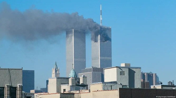 The twin towers stand tall against a bright blue sky, with other buildings visible in front of them. Black smoke billows from a huge hole in the top of the tower on the right.