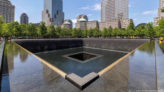 A big, square, marble rim around a sunken area, photographed from a striking angle. Behind it is a row of trees, and behind that several New York skyscrapers.