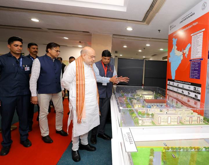 A group of individuals observes a detailed architectural model at a presentation, featuring a large map of India and an informational display on the wall.