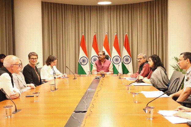 A meeting scene with multiple people seated around a long conference table, featuring the Indian national flags in the background. The central figure is speaking, with others attentively listening.