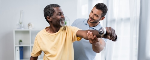 A patient holds a dumbell with one arm, being assisted by physical therapist