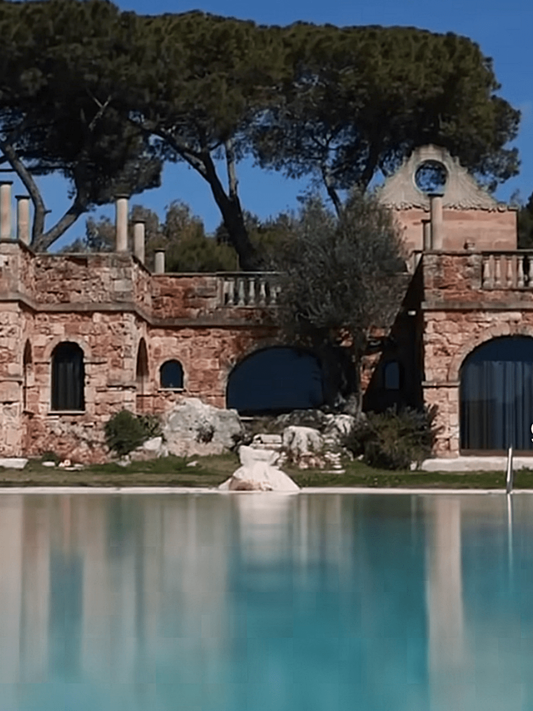 Spiegelglatte Wasseroberfläche vor einer rustikalen Steinarchitektur mit Pinien im Hintergrund.