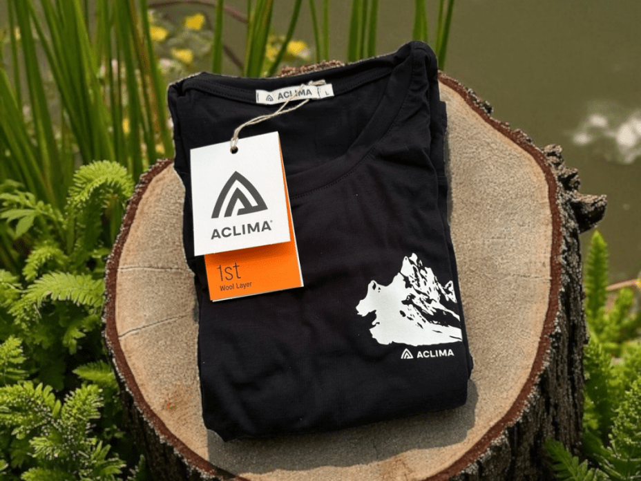 Aclima Classic Tee in Schwarz, gefaltet auf einem Baumstumpf mit sichtbarem Berg-Print und Aclima-Etikett.