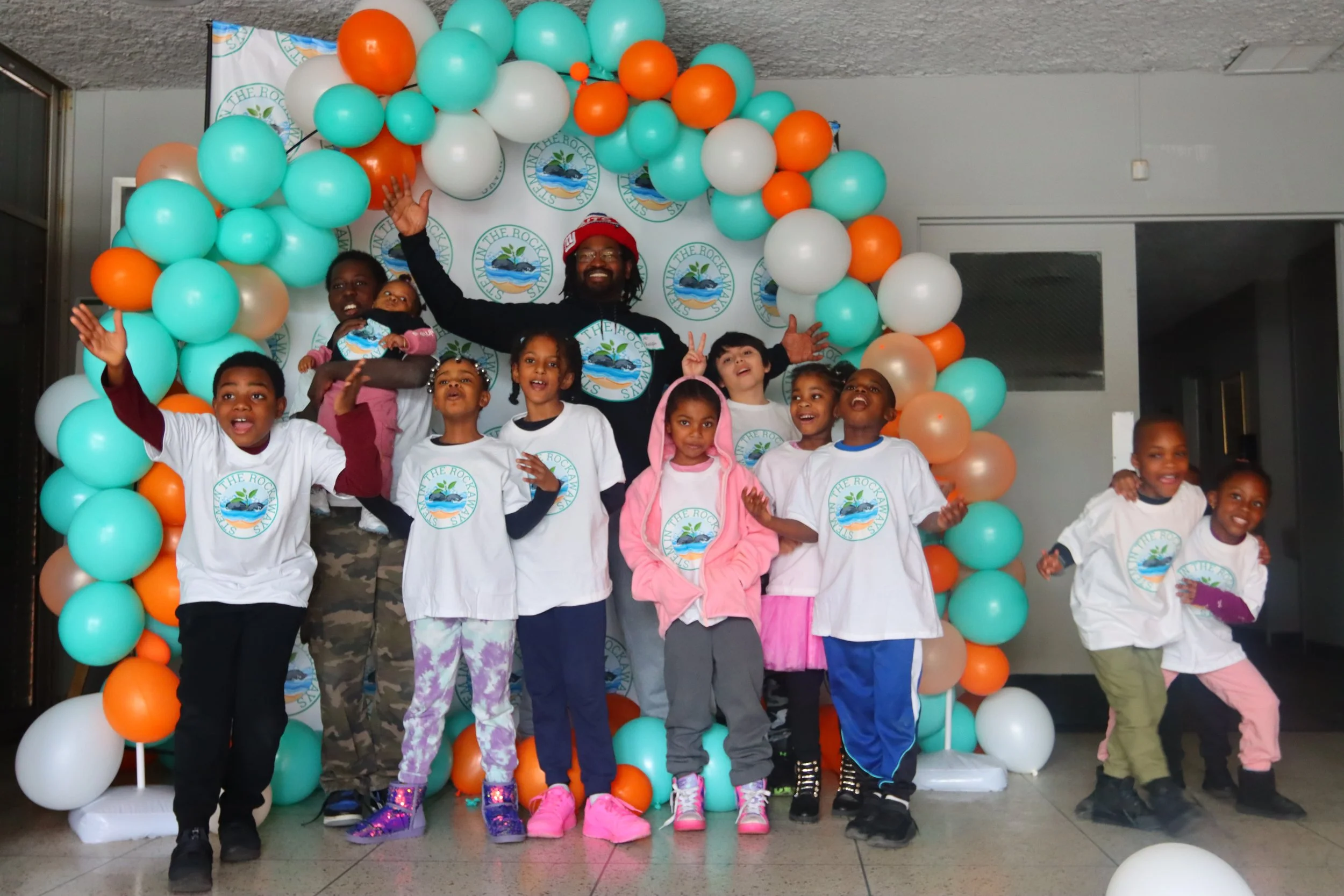 Smiling young learners in STEM In The Rockaways T-shirts celebrating in front of a teal and orange balloon arch