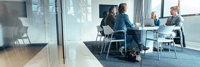 A group of business people engage in a tele-conference at a large table inside an office with glass walls.