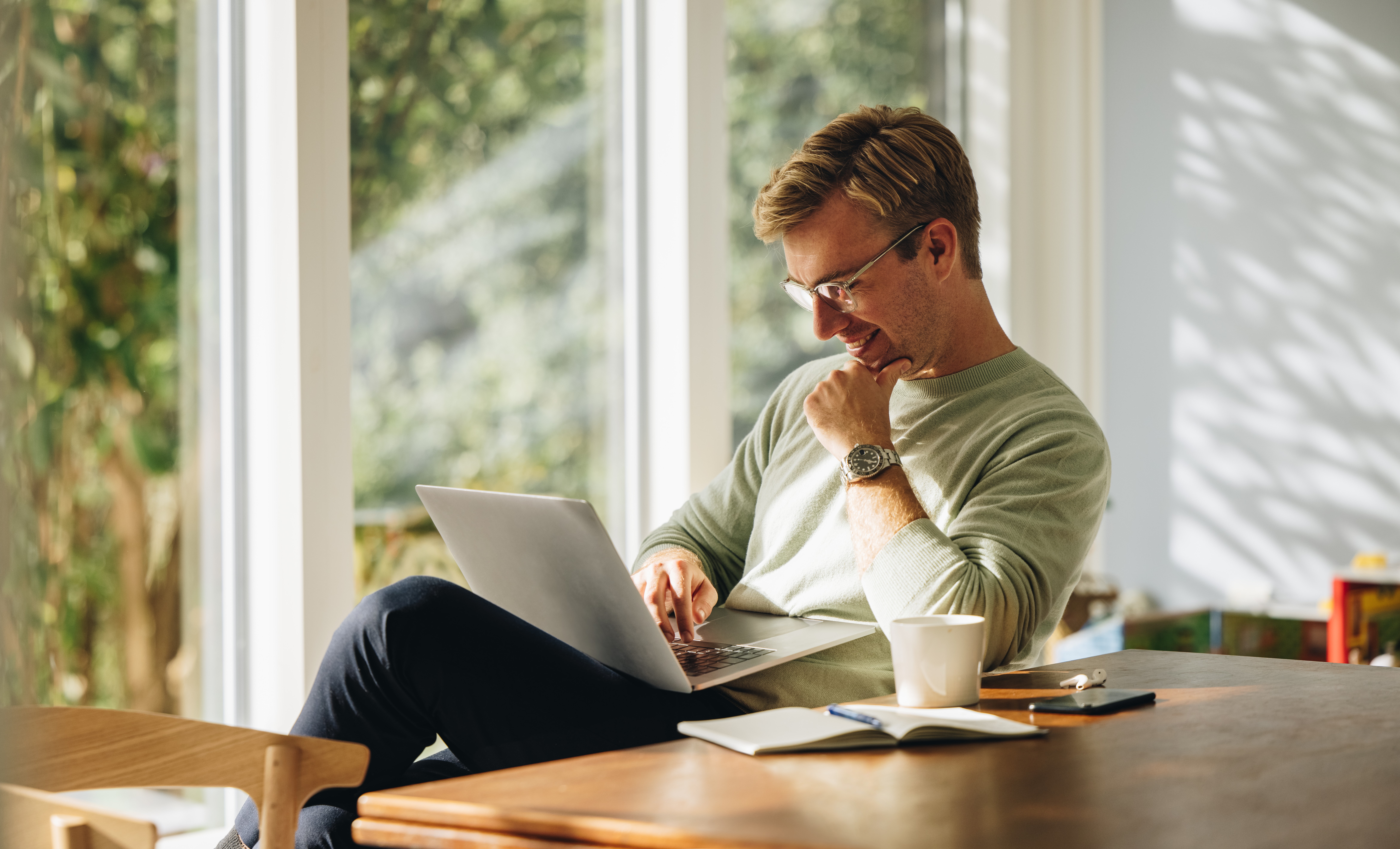 A man wearing glasses and a light sweater smiles as he works on a laptop at a wooden table. A notebook, pen, and coffee mug sit beside him, with a large window and greenery visible in the background.