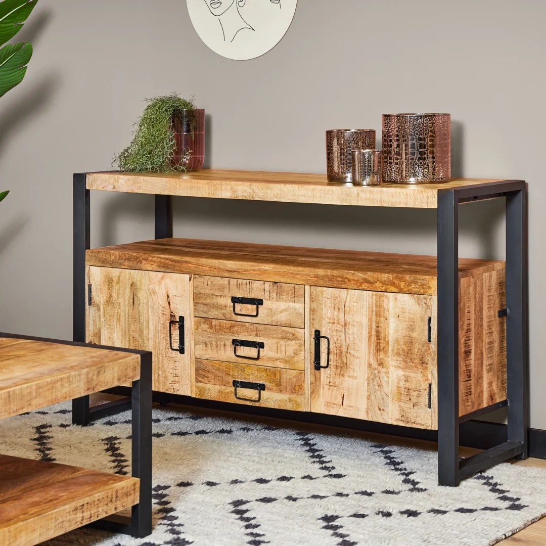 De Dressoir Boaz 135 cm is een modern houten dressoir met een zwart metalen frame, drie centrale lades en twee zijkasten. Decoratieve items rusten bovenop, terwijl het staat op een gedessineerd tapijt tegen een beige muur.