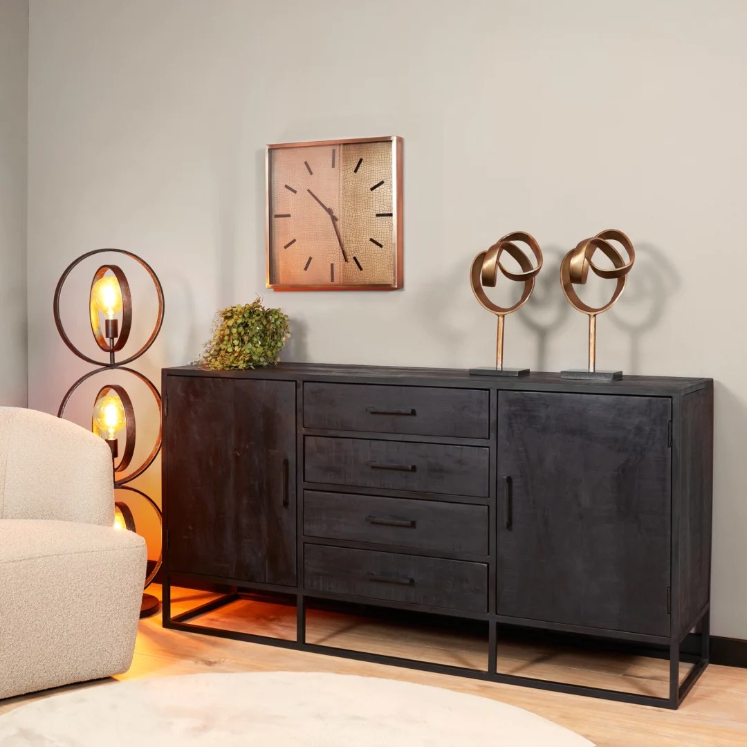Een moderne woonkamer met de Dressoir Denver Black 165 cm in mangohout en staal, abstracte gouden sculpturen, een wandklok, een unieke vloerlamp met drie verlichte lampen en een gedeeltelijk zichtbare beige fauteuil aan de linkerkant.