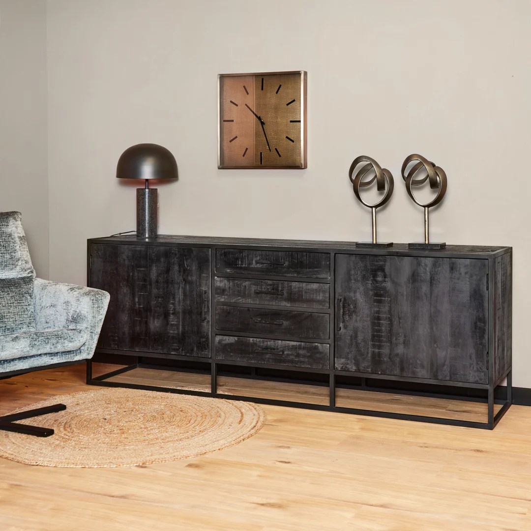 Een moderne woonkamer met het Dressoir Denver Black 210 cm dressoir in mangohout en staal, twee abstracte sculpturen, een zwarte tafellamp, een rond geweven vloerkleed, een metalen wandklok en een deel van een grijs gestoffeerde stoel op een licht houten vloer.