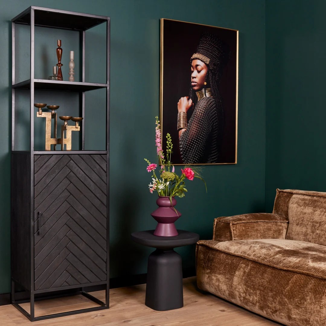 Een moderne woonkamer met donkergroene muren, een bruin fluwelen bank, de Boekenkast New York mangohout en staal met decor, een zwart bijzettafeltje met een paarse vaas met kleurrijke bloemen en een ingelijst portret van een vrouw in traditionele kleding.