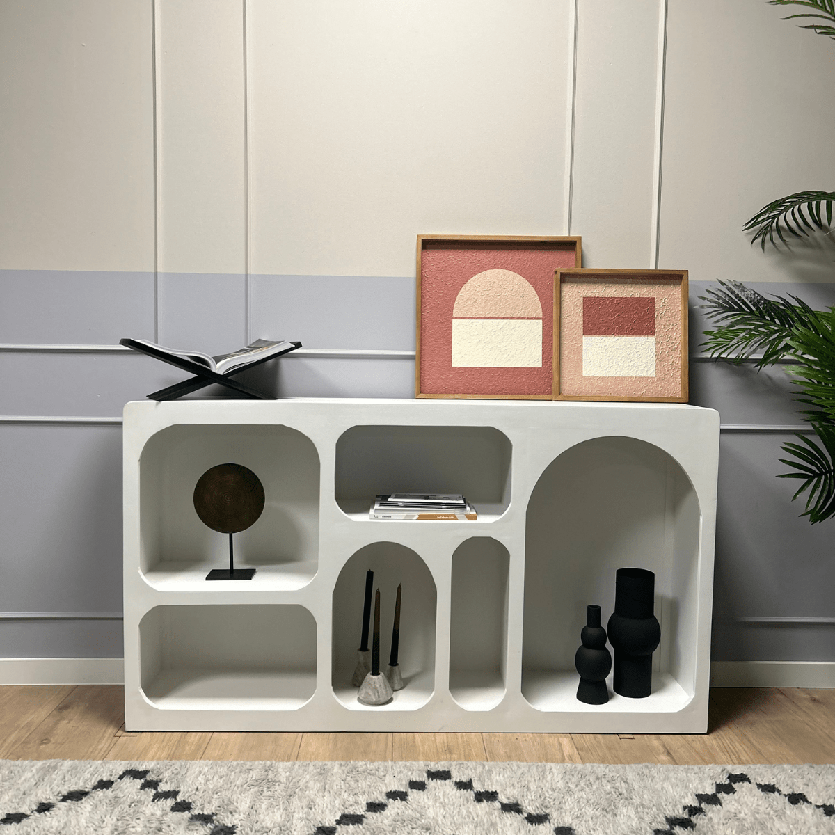 De Dressoir Caya Wit, versierd met abstracte kunst in houten frames, zwarte vazen, kwasten, een rond beeldhouwwerk en een open boek, staat tegen een licht gepaneelde muur naast een groene plant.