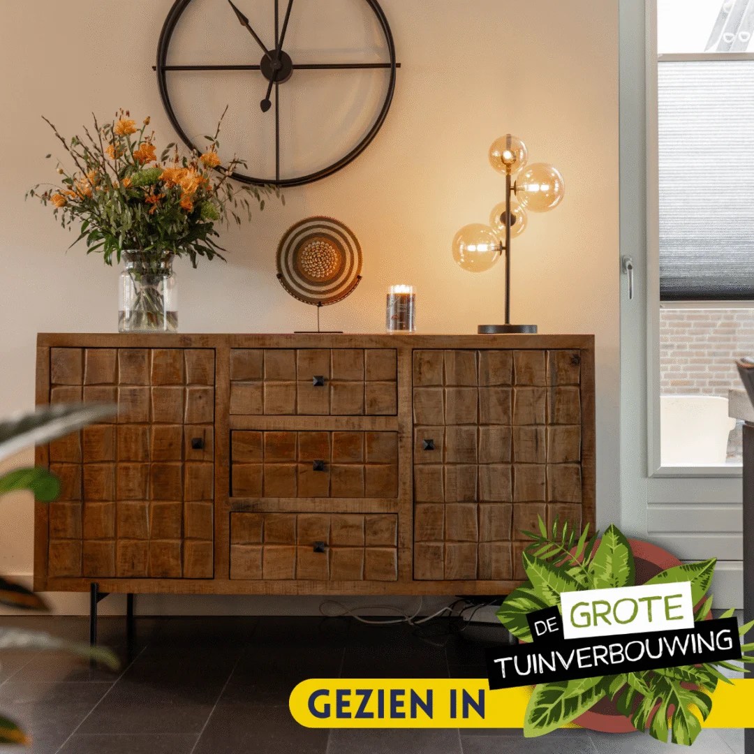 De Dressoir Brandy | 160 cm, met een geruit patroon, staat tegen de muur met een vaas, bord en lamp erop. Een grote klok hangt erboven. Overlay tekst: "Gezien in De Grote Tuinverbouwing.