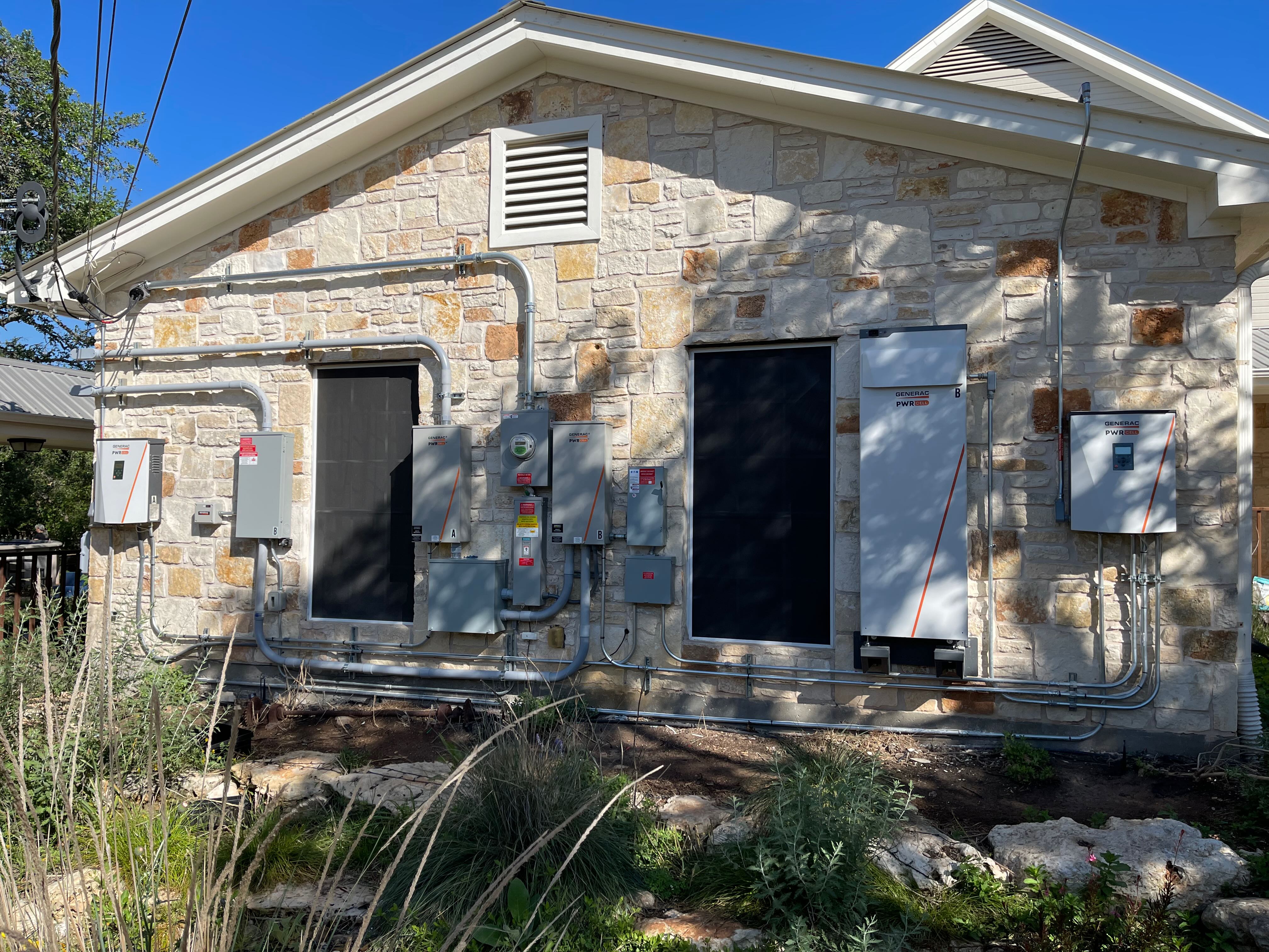 Outside wall of a house adorned with multiple boxes of solar power equipment connected by conduit, including inverters, transfer switches, and batteries