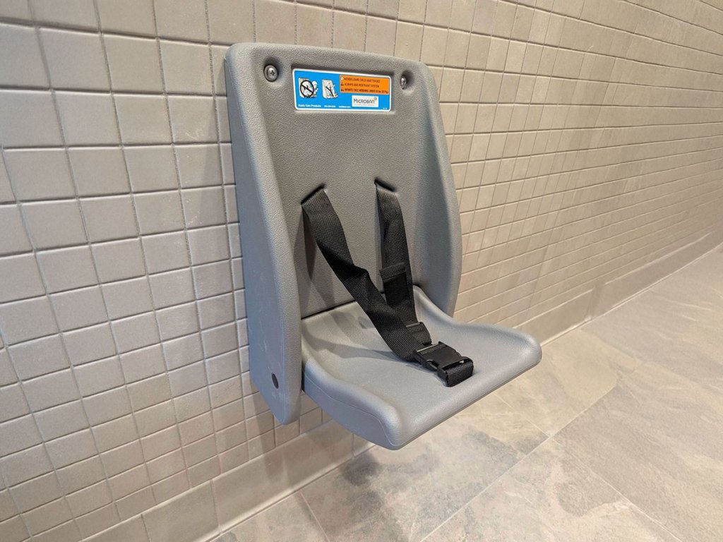 A gray, wall-mounted Koala Kare child safety seat shown in the open position against a tiled wall. The seat has a black harness with a buckle to secure a child.