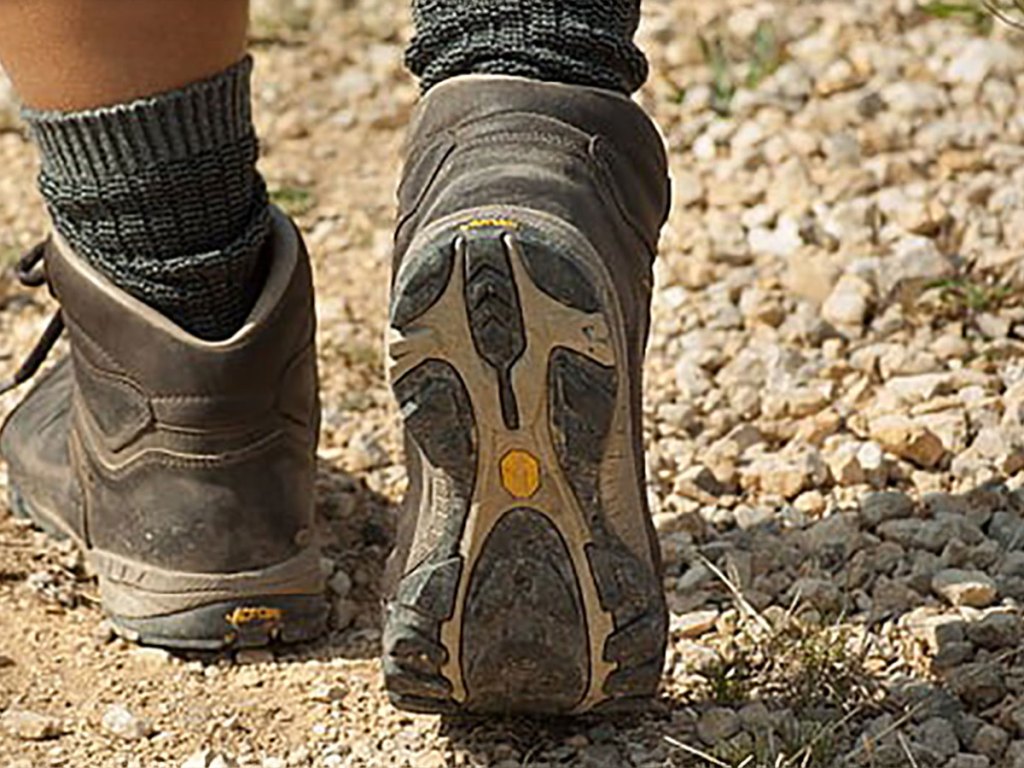 A close-up from behind of a person wearing brown hiking boots and grey wool socks, walking on a rocky, dirt trail.